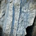 Ein großer, grob behauener Block aus hellem Naturstein mit markanten, senkrecht verlaufenden blauen Streifen. die Oberfläche ist rau und uneben, mit sichtbaren Bruchkanten und natürlichen Adern. Der Stein zeugt eine Mischung aus hellgrauen und bläulichen Farbtönen. Es handelt sich um einen Quarzit