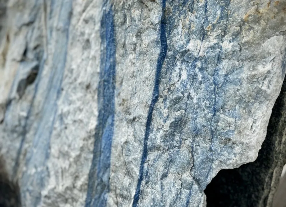 Ein großer, grob behauener Block aus hellem Naturstein mit markanten, senkrecht verlaufenden blauen Streifen. die Oberfläche ist rau und uneben, mit sichtbaren Bruchkanten und natürlichen Adern. Der Stein zeugt eine Mischung aus hellgrauen und bläulichen Farbtönen. Es handelt sich um einen Quarzit