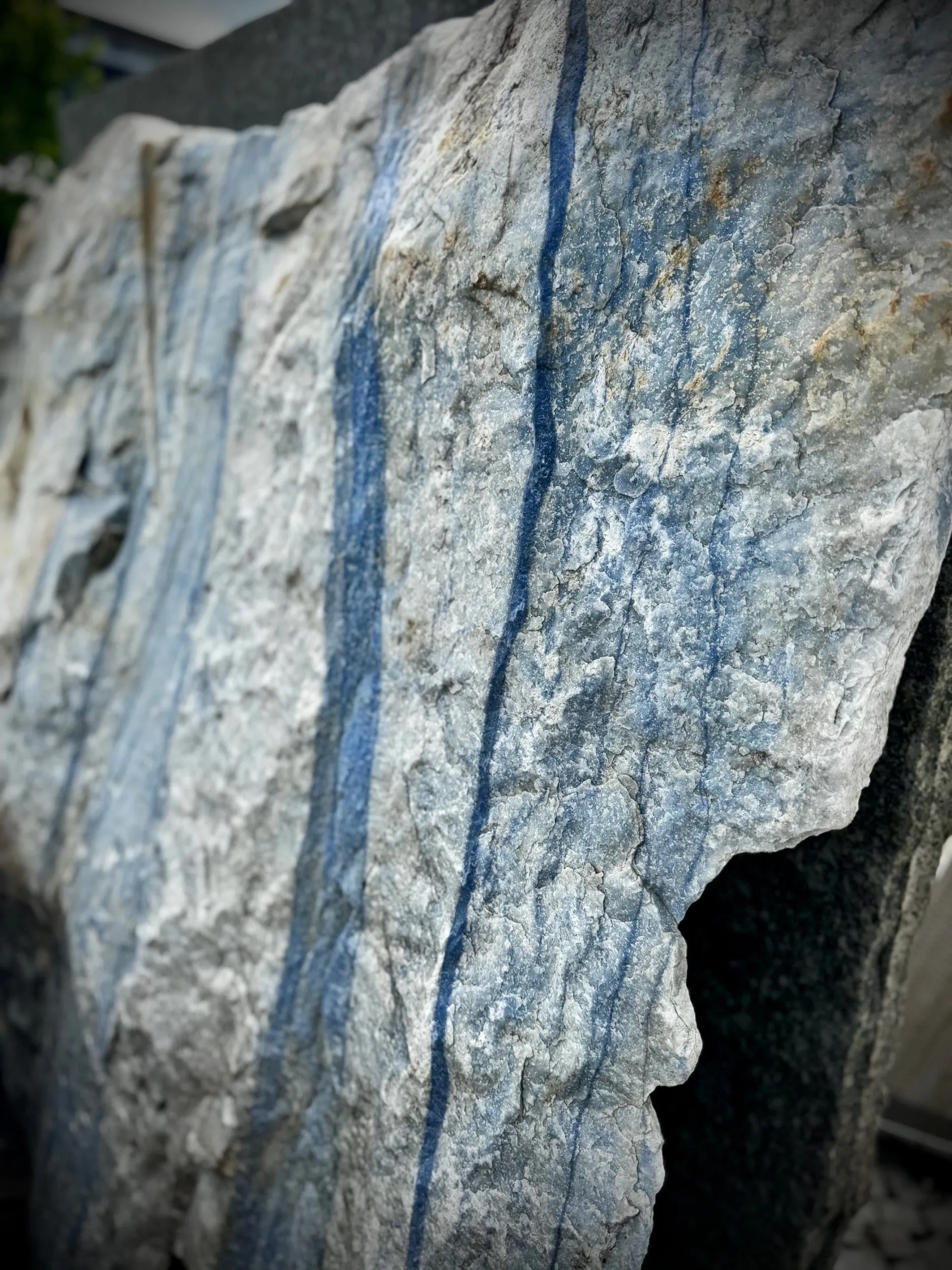 Ein großer, grob behauener Block aus hellem Naturstein mit markanten, senkrecht verlaufenden blauen Streifen. die Oberfläche ist rau und uneben, mit sichtbaren Bruchkanten und natürlichen Adern. Der Stein zeugt eine Mischung aus hellgrauen und bläulichen Farbtönen. Es handelt sich um einen Quarzit