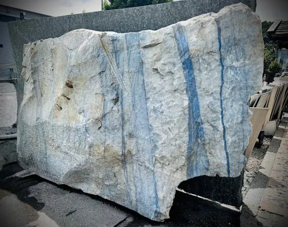 Ein großer, grob behauener Block aus hellem Naturstein mit markanten, senkrecht verlaufenden blauen Streifen. die Oberfläche ist rau und uneben, mit sichtbaren Bruchkanten und natürlichen Adern. Der Stein zeugt eine Mischung aus hellgrauen und bläulichen Farbtönen. Es handelt sich um einen Quarzit